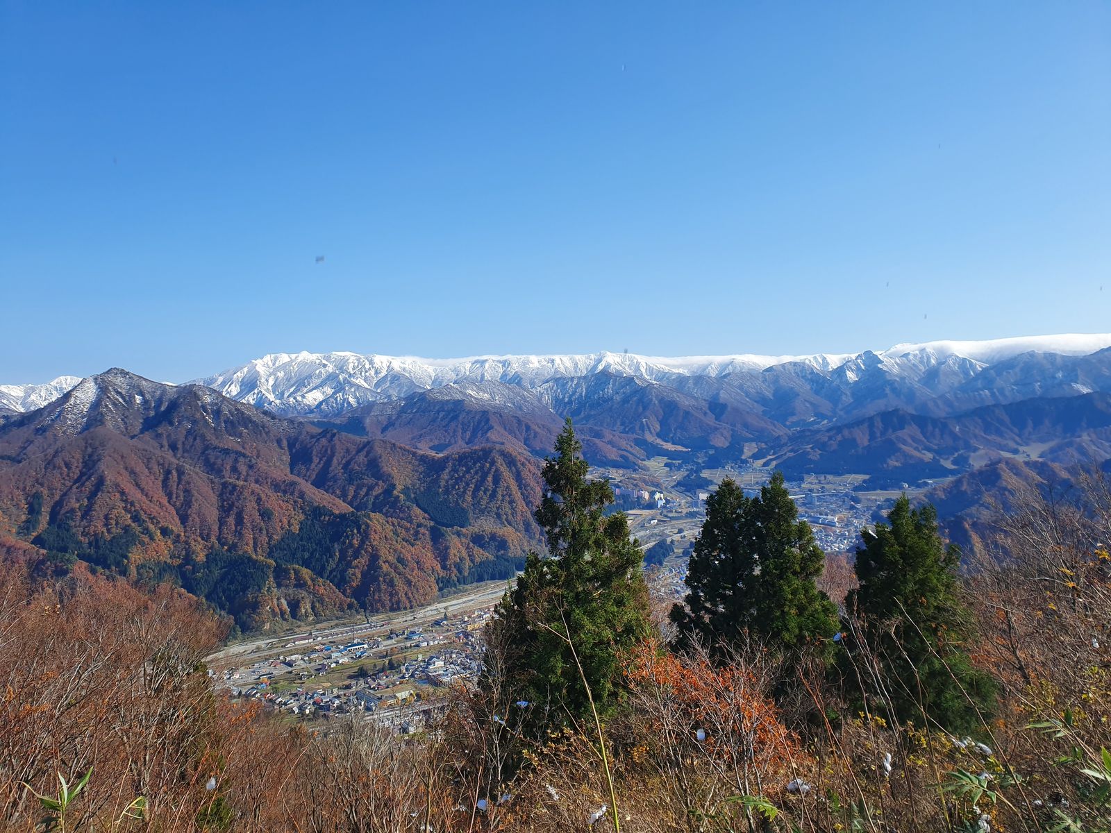 紅葉の山あいに広がる越後湯沢の市街地を高台から俯瞰した写真。中央にリゾートマンション群と上越新幹線の高架が見える