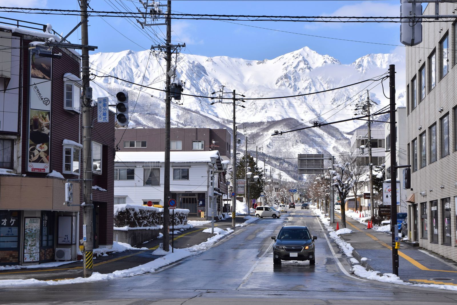 冬の白馬村中心部。除雪された県道の正面に雪をまとった北アルプス白馬三山がそびえ、道路沿いに宿泊施設やスポーツ用品店が並ぶ
